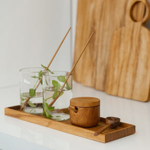 A wooden tray holding two clear glasses with water and mint leaves and two long wooden stirring spoons, a round wooden jar with a lid, and a small wooden spoon, set on a white surface with a blurred wooden cutting board in the background.