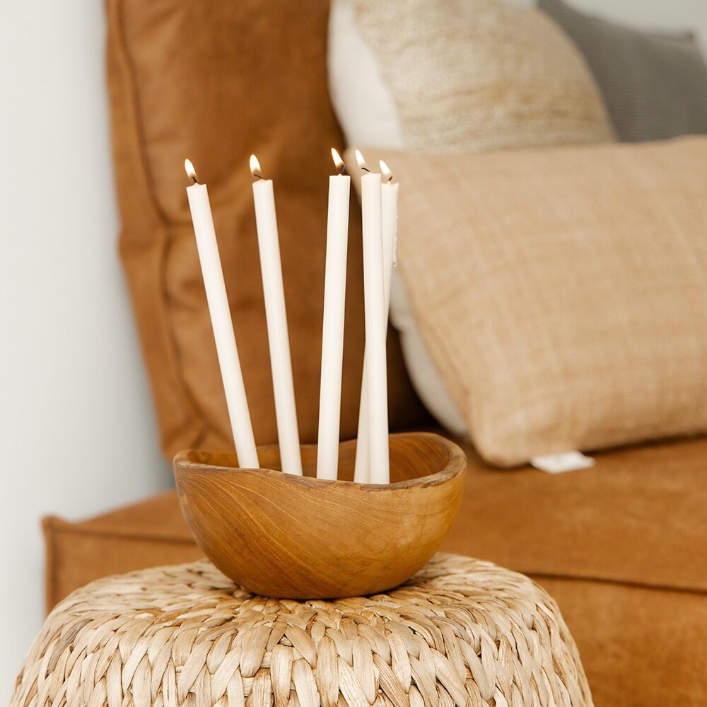 Five white thin candles with lit flames, standing in a small wooden bowl placed on a woven side table in a cozy living room setting.
