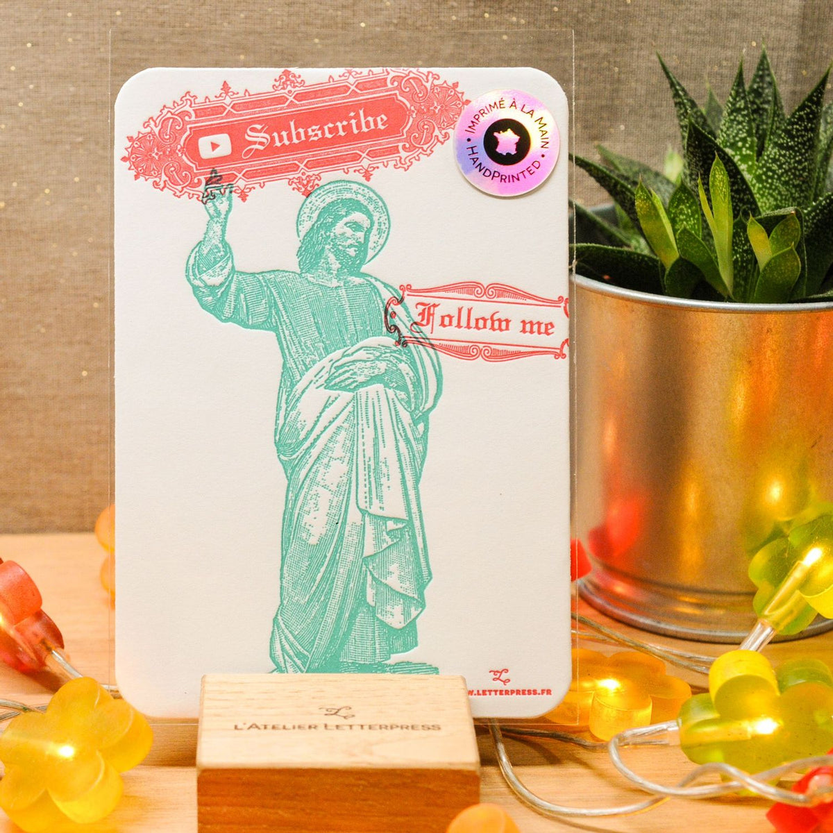 A rectangular white art print card with a teal illustration of a robed religious figure and decorative red text reading 'Subscribe' and 'Follow me,' styled on a tabletop with a small wooden block, string lights, and a potted succulent in the background.