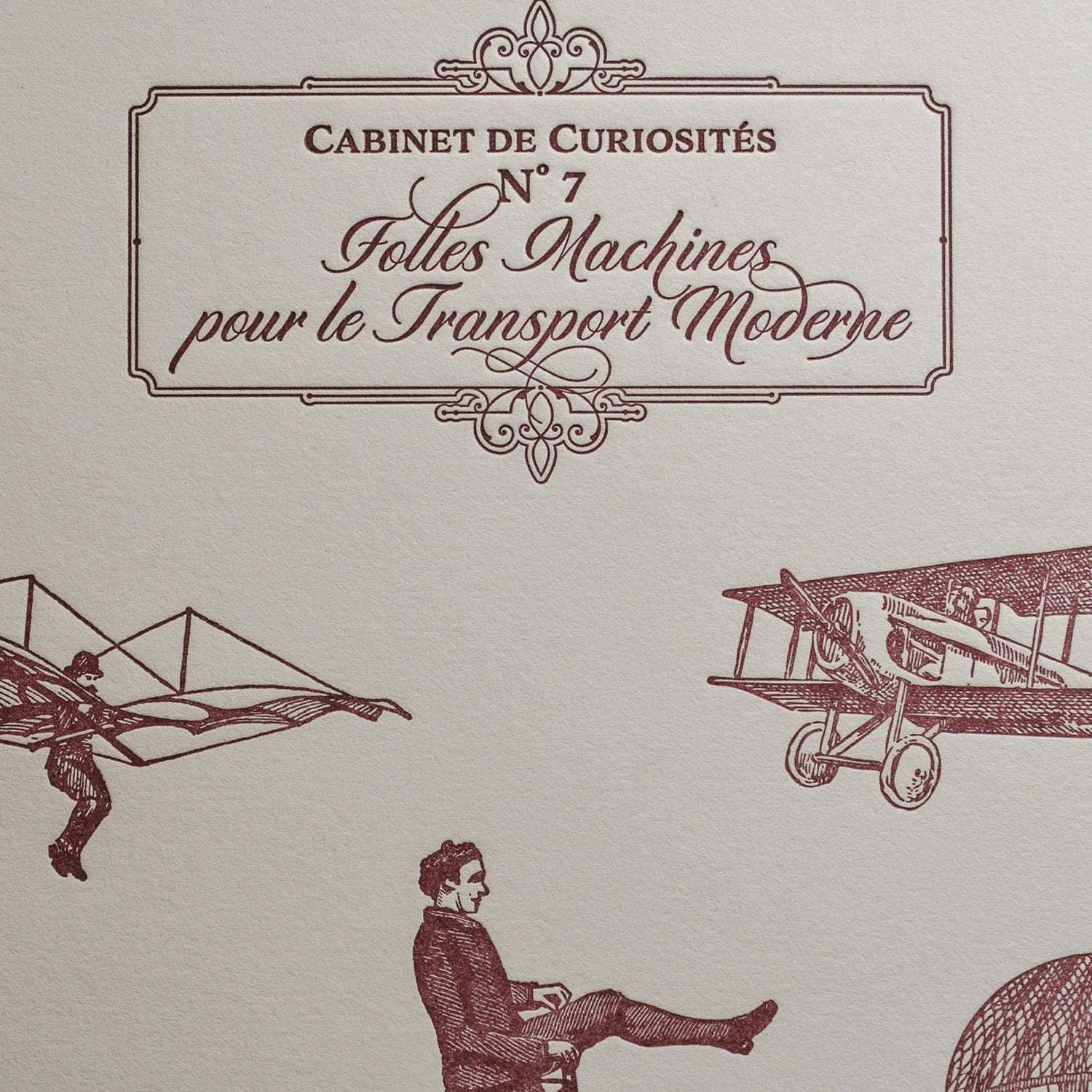 A close up of vintage-style illustrated art print with French text in an ornate frame, featuring burgundy drawings of an airplane, a winged figure, and a seated man on an old bicycle on off-white cotton paper.