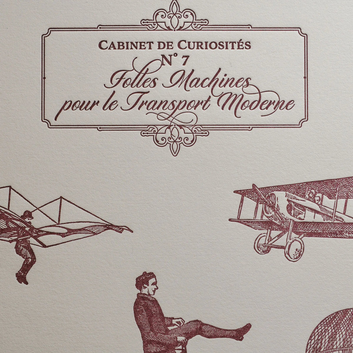 A close up of vintage-style illustrated art print with French text in an ornate frame, featuring burgundy drawings of an airplane, a winged figure, and a seated man on an old bicycle on off-white cotton paper.