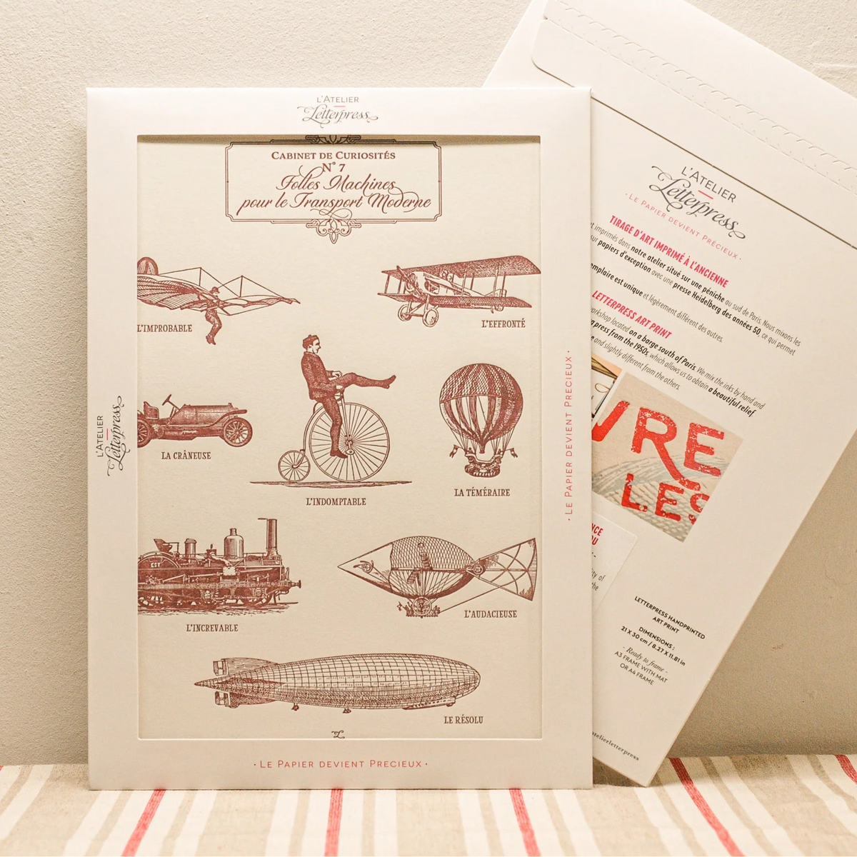 A vintage-style art print featuring burgundy illustrations of early automobiles and transportation inventions, displayed in a cardboard envelope with another showing the back side behind it on a striped fabric surface against a beige wall.