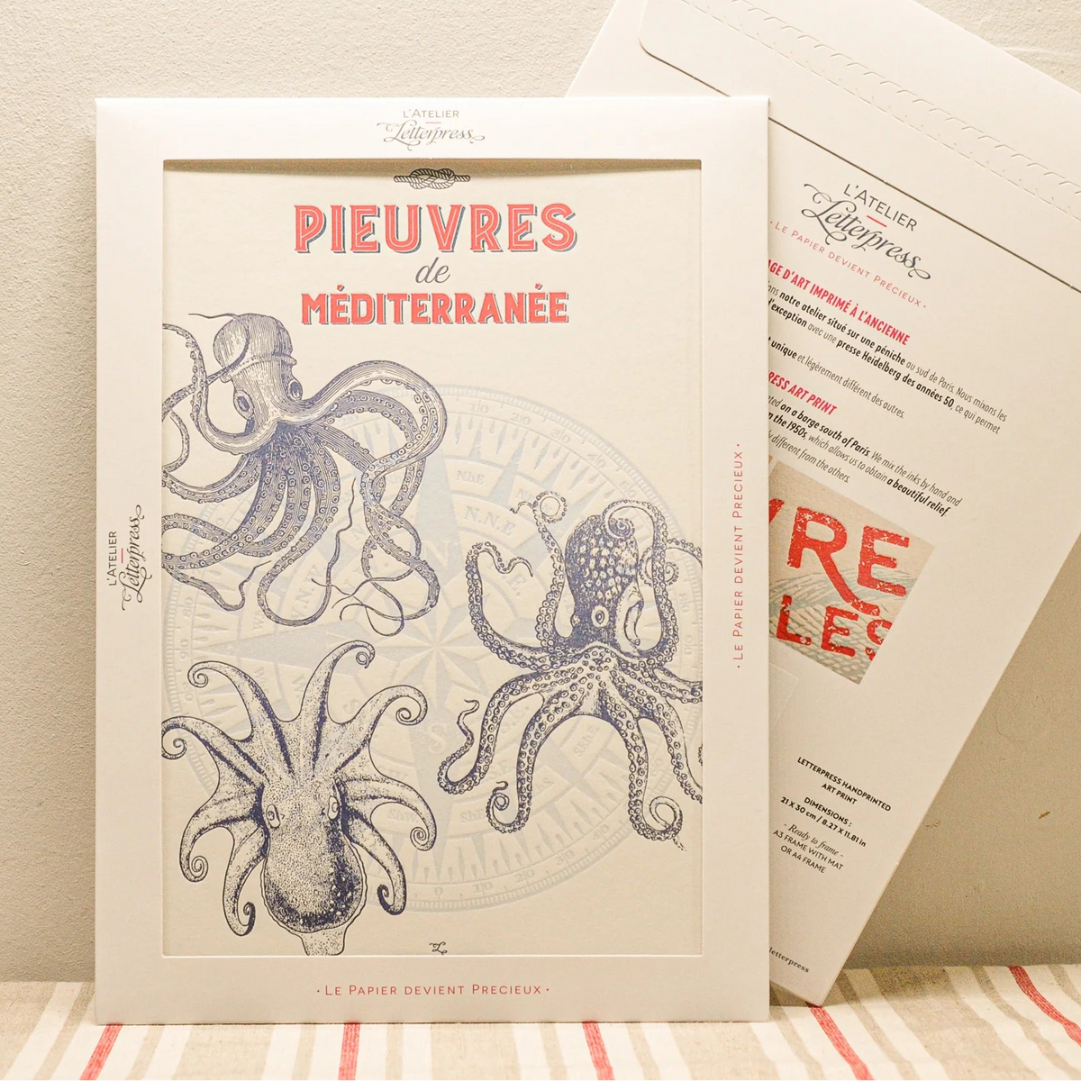 A vintage-style art print featuring dark blue illustrations of octopuses, displayed in a cardboard envelope with another showing the back side behind it on a striped fabric surface against a beige wall.