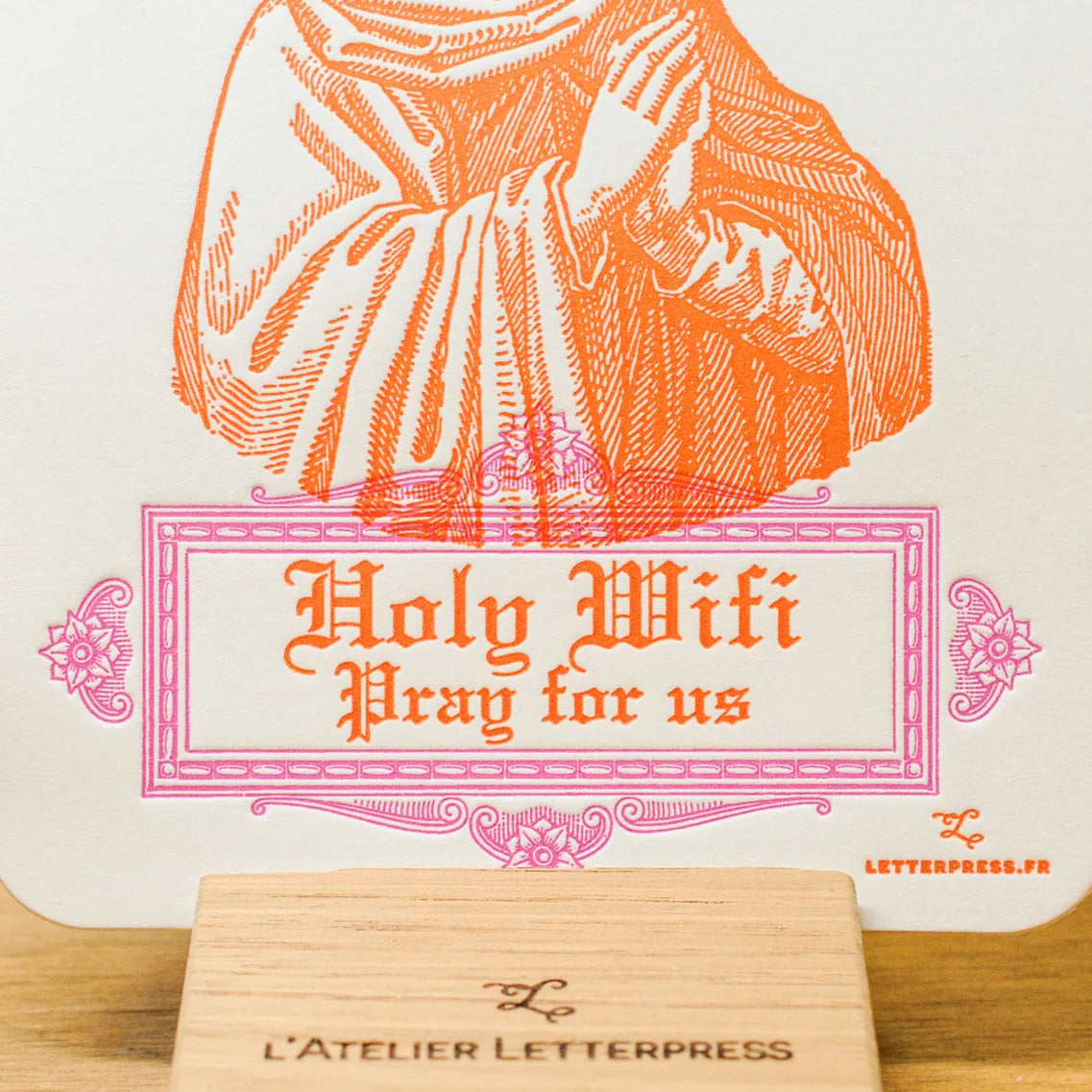 A close up of a white paper art print card with an orange line-art illustration of a robed figure and the text 'Holy Wifi Pray for us' in orange, displayed on a small wooden stand.