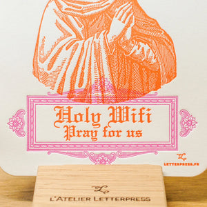 A close up of a white paper art print card with an orange line-art illustration of a robed figure and the text 'Holy Wifi Pray for us' in orange, displayed on a small wooden stand.