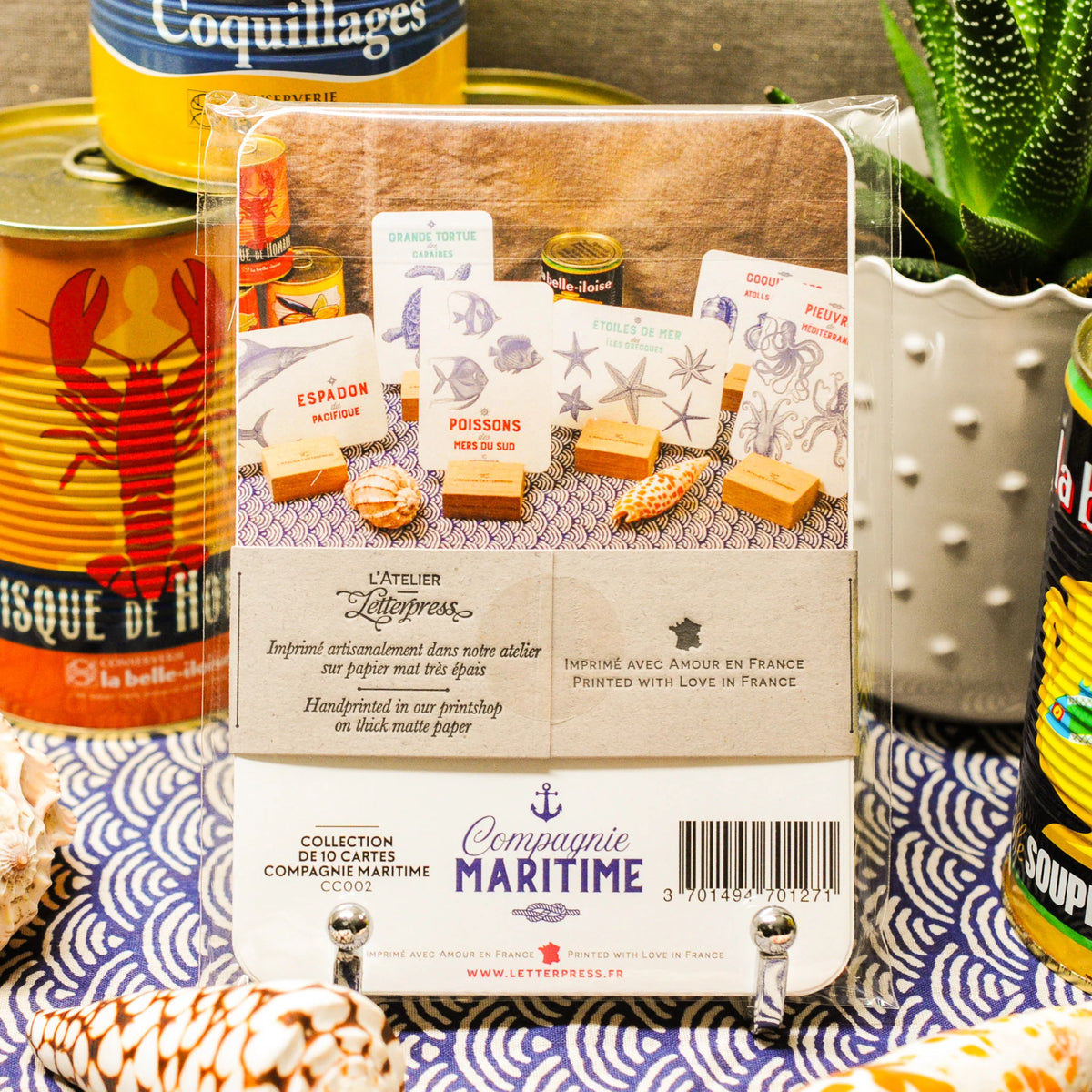 Packaged set of 10 Maritime sea creatures art print cards bound together with a recycled brown paper strip shown from the back on a surface, surrounded by canned seafood, shells, and a succulent in a styled setting.