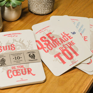 A set of French text set of 10 anatomy art print cards spread out and displayed on a wooden tabletop. The cards are coloured with red and gray text and illustrations, and a paper band wraps one card.
