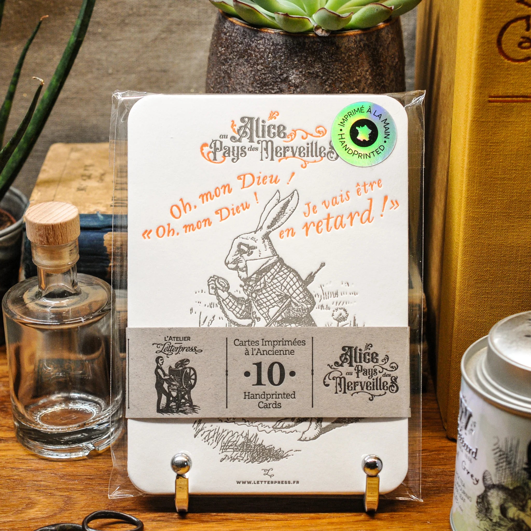 Packaged set of 10 Alice in Wonderland art print cards bound together with a recycled brown paper strip on a wooden surface with decorative items.