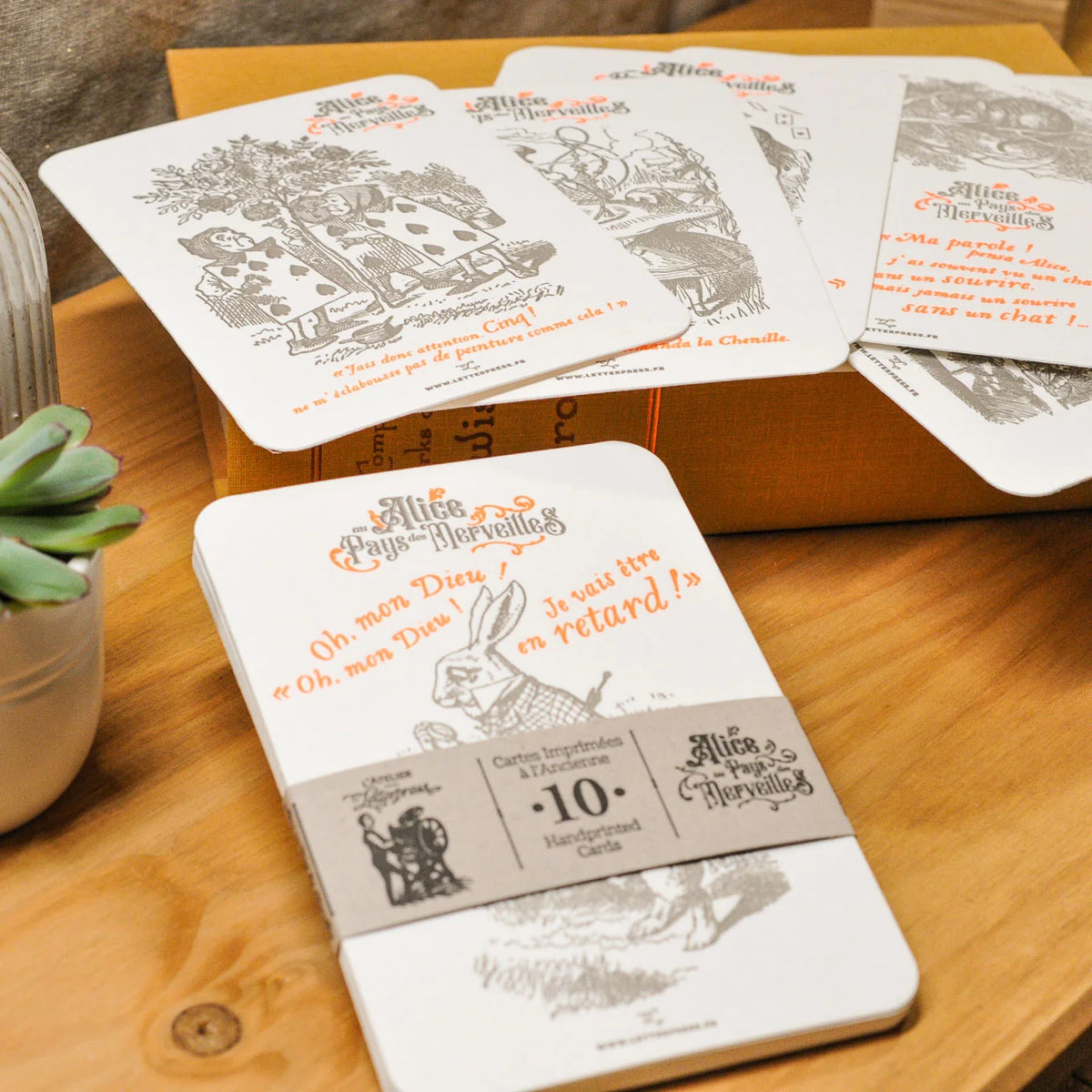 A set of French text set of 10 Alice in Wonderland art print cards spread out and displayed on a wooden tabletop. The cards are coloured with red and gray text and illustrations, and a paper band wraps one card.