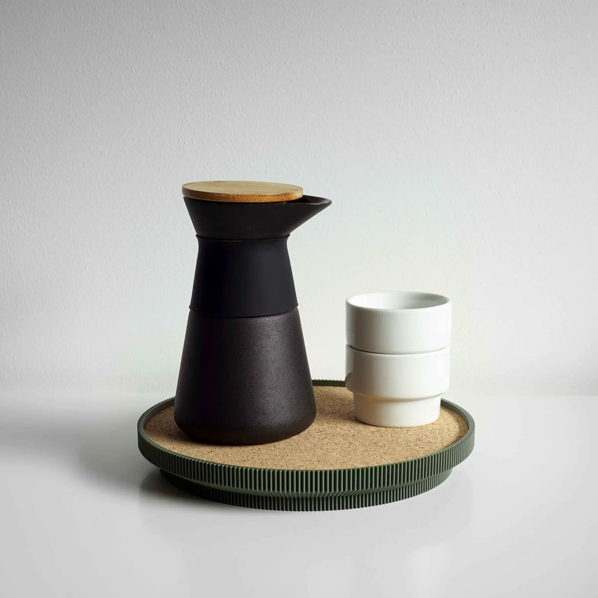A matte black ceramic carafe with a light wood lid sits on a round green cork topped tray next to two stacked white ceramic cups, set against a neutral light gray background.
