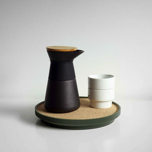 A matte black ceramic carafe with a light wood lid sits on a round green cork topped tray next to two stacked white ceramic cups, set against a neutral light gray background.