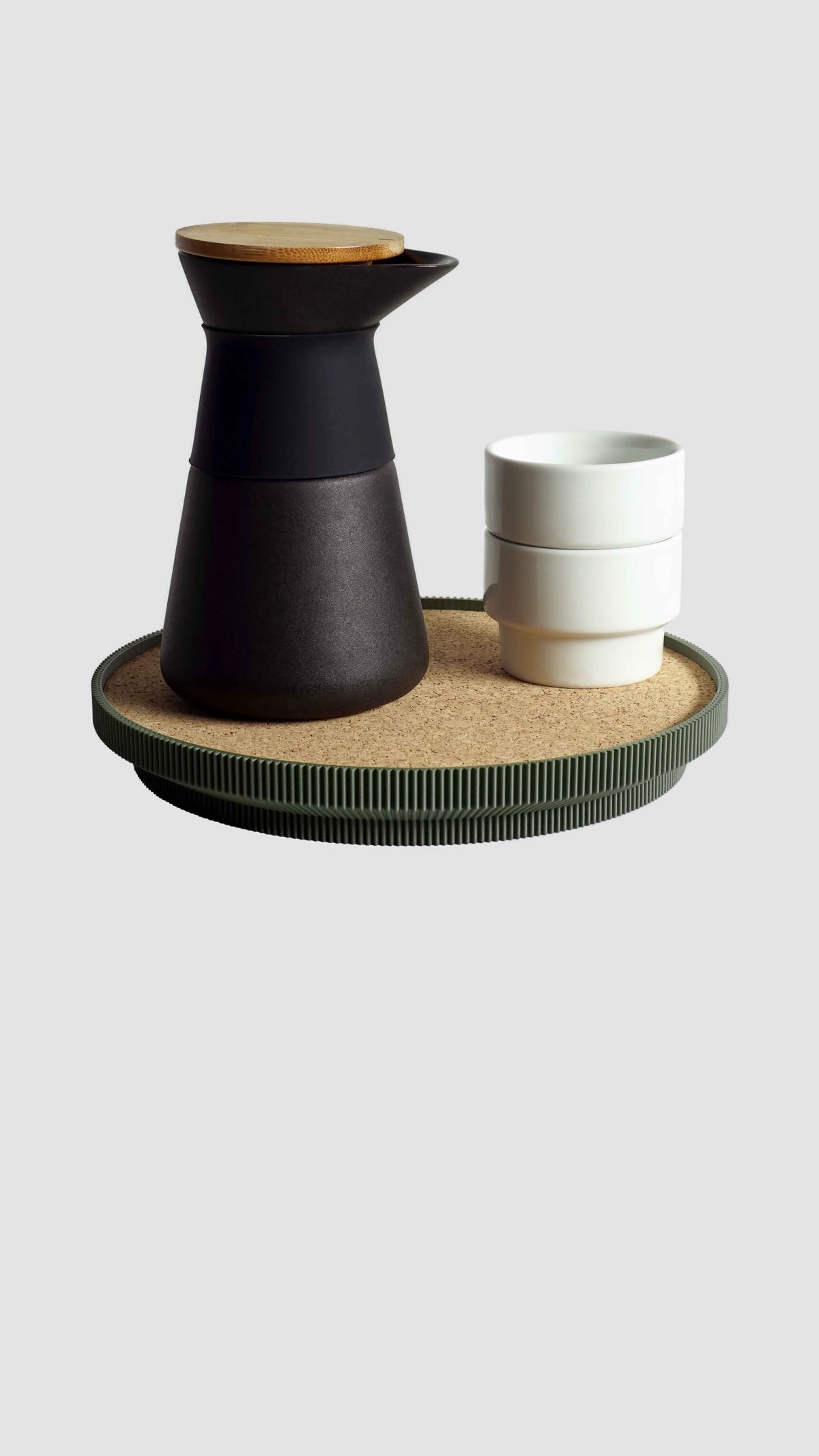 A modern coffee carafe set in matte black with a light wood lid, served on a round ribbed tray with a cork top next to two stacked white cups, against a solid light gray background.