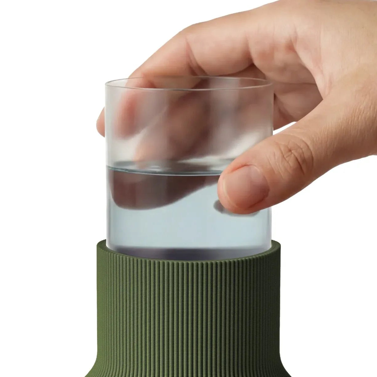 A close-up of a hand holding a clear plastic insert with water inside it, above a ribbed olive green bottle vase on a white background.