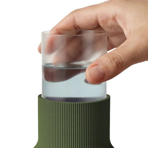 A close-up of a hand holding a clear plastic insert with water inside it, above a ribbed olive green bottle vase on a white background.