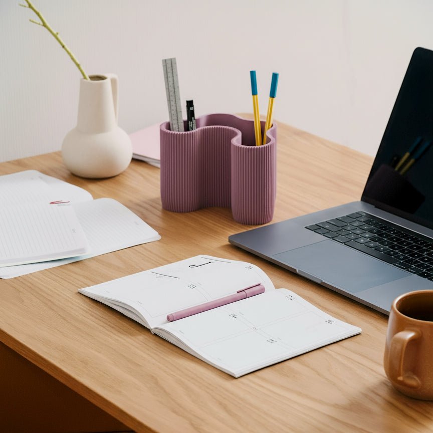 A modern four-compartment dusky-purple desk organiser with pens and a ruler inside it, on a wooden work desk with a laptop, notebook and vase.