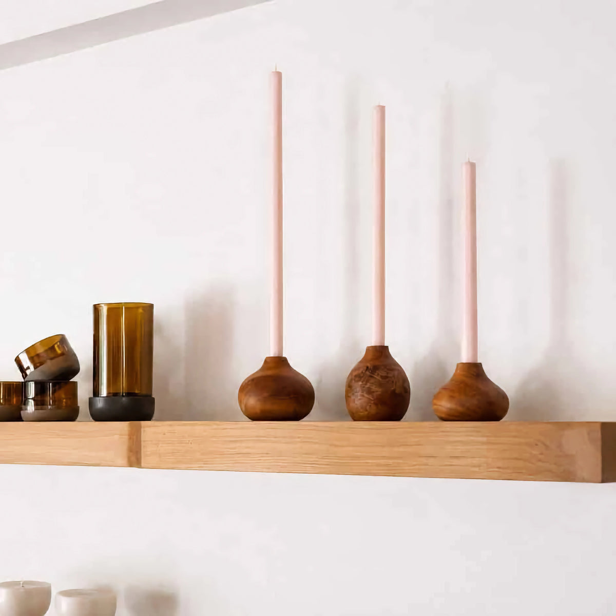 Three tall blush pink candles in rounded wooden holders displayed on a light oak floating shelf against a white wall.