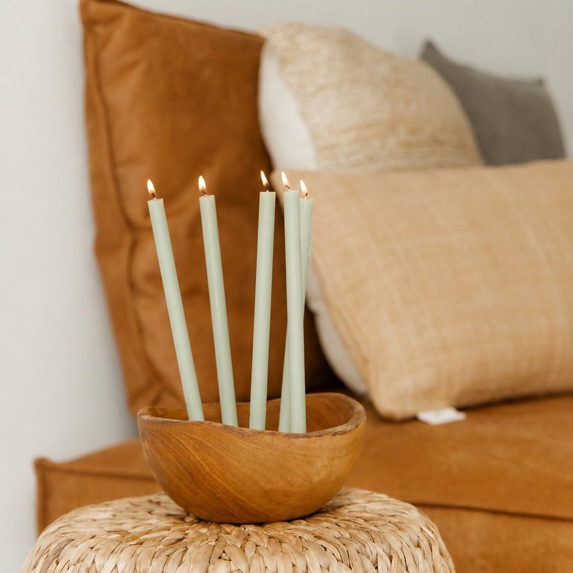 Five light green thin candles with lit flames, standing in a small wooden bowl placed on a woven side table in a cozy living room setting.