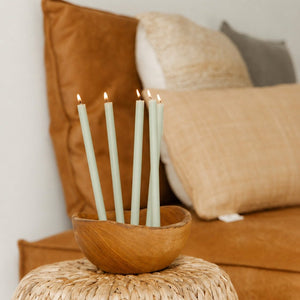 Five light green thin candles with lit flames, standing in a small wooden bowl placed on a woven side table in a cozy living room setting.