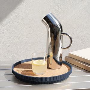 A polished stainless steel pitcher with a curved spout and side handle sits on a navy blue round cork topped tray beside a small clear glass of lemon water on a light gray slatted table.