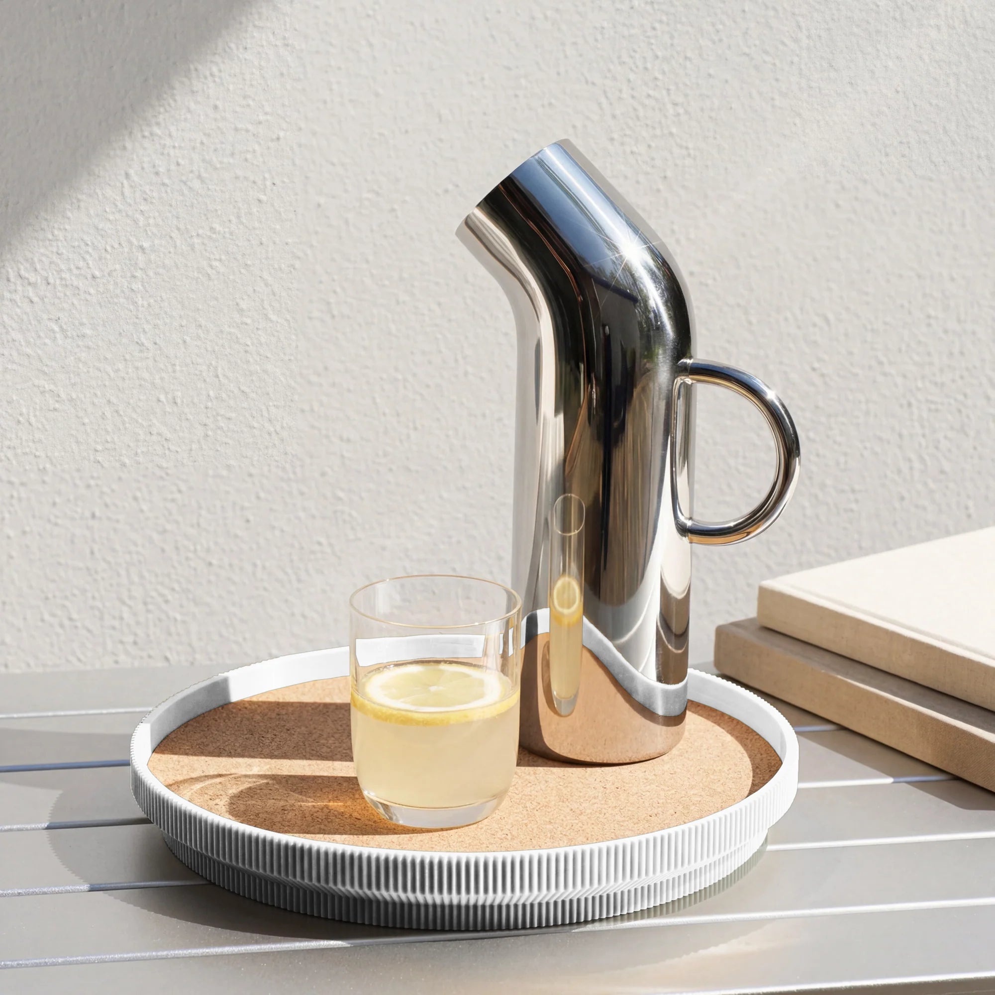A polished stainless steel pitcher with a curved spout and side handle sits on a white round cork topped tray beside a small clear glass of lemon water on a light gray slatted table.