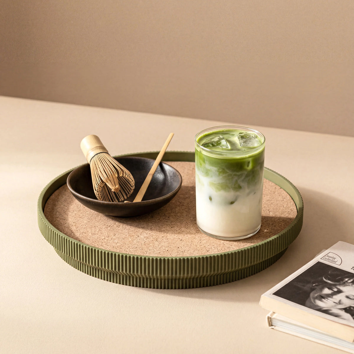 Glass of iced matcha green tea on a green rimmed cork tray with a whisk and bowl against a beige background