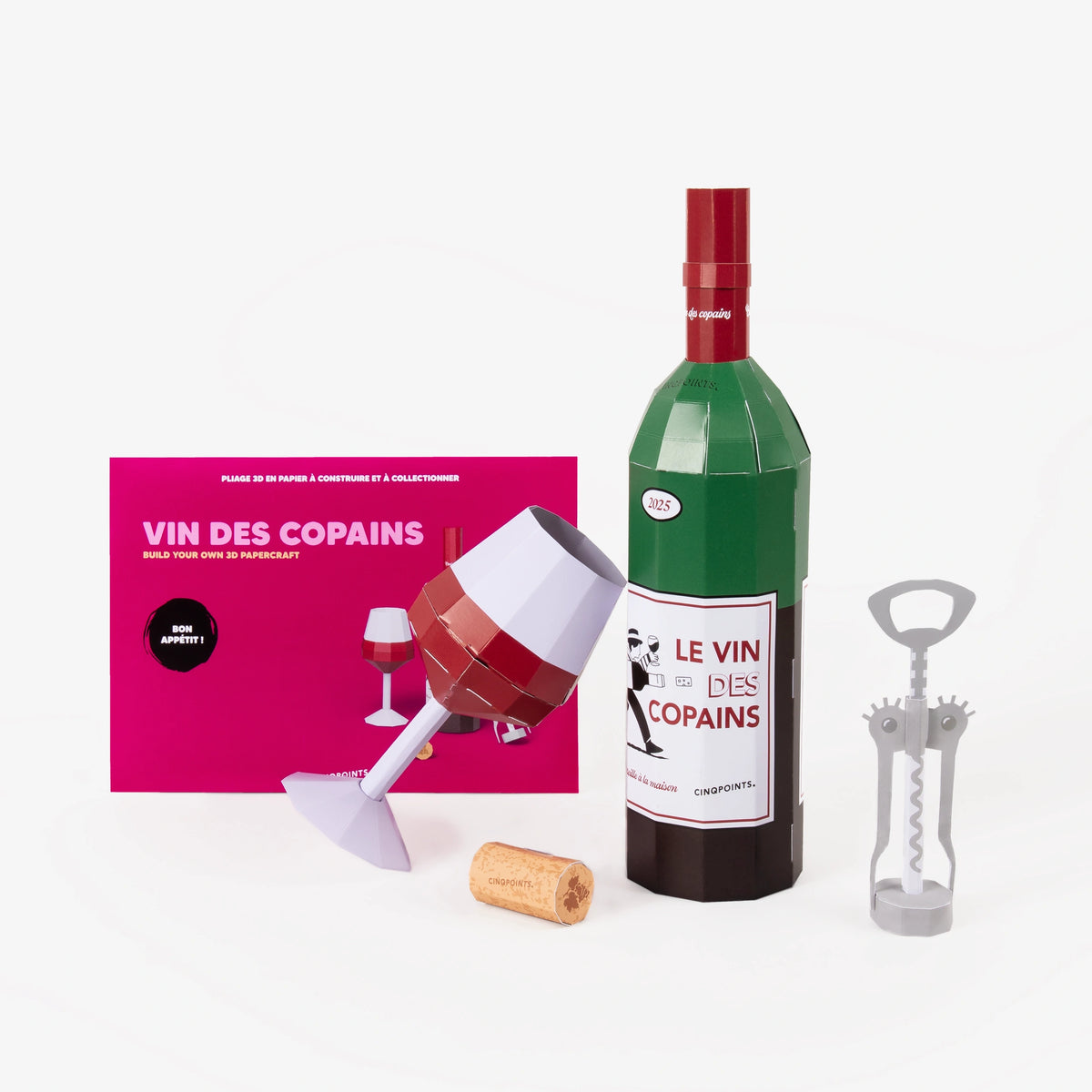 A pink cardboard envelope for a wine paper model scene set in the background, in front paper models of a wine bottle, wine glass, corkscrew, and cork on the front, photographed on a clean white background.