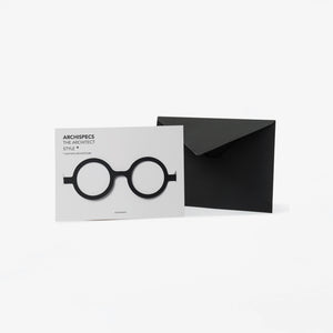 A minimalist architectural card set featuring a white card with black cut out cardboard circular eyeglass, shown next to a black envelope on a light gray background.