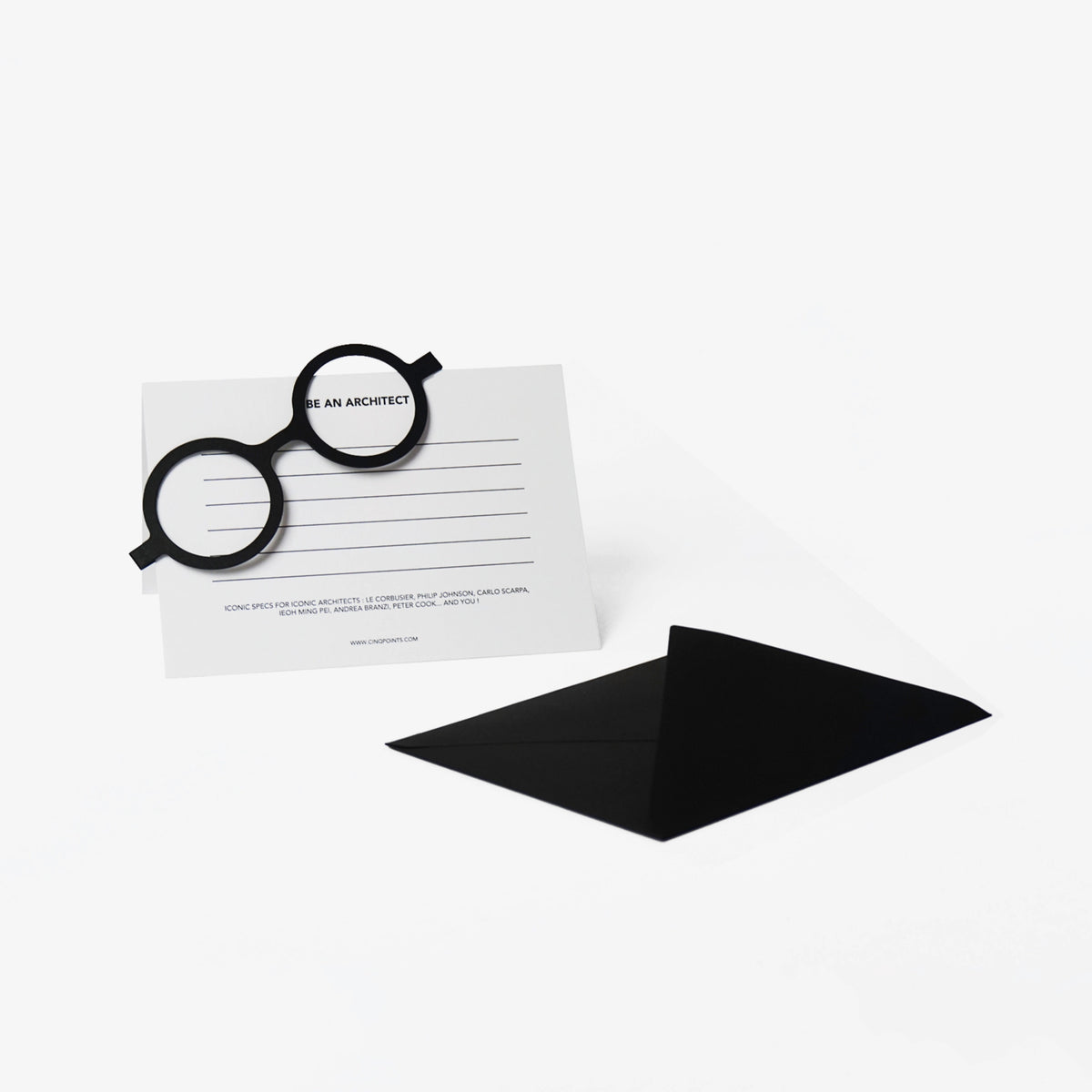 A minimalist architectural card set featuring black cut out cardboard circular eyeglass resting on a white lined card with the text 'BE AN ARCHITECT', next to a black envelope on a clean white background.