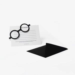 A minimalist architectural card set featuring black cut out cardboard circular eyeglass resting on a white lined card with the text 'BE AN ARCHITECT', next to a black envelope on a clean white background.