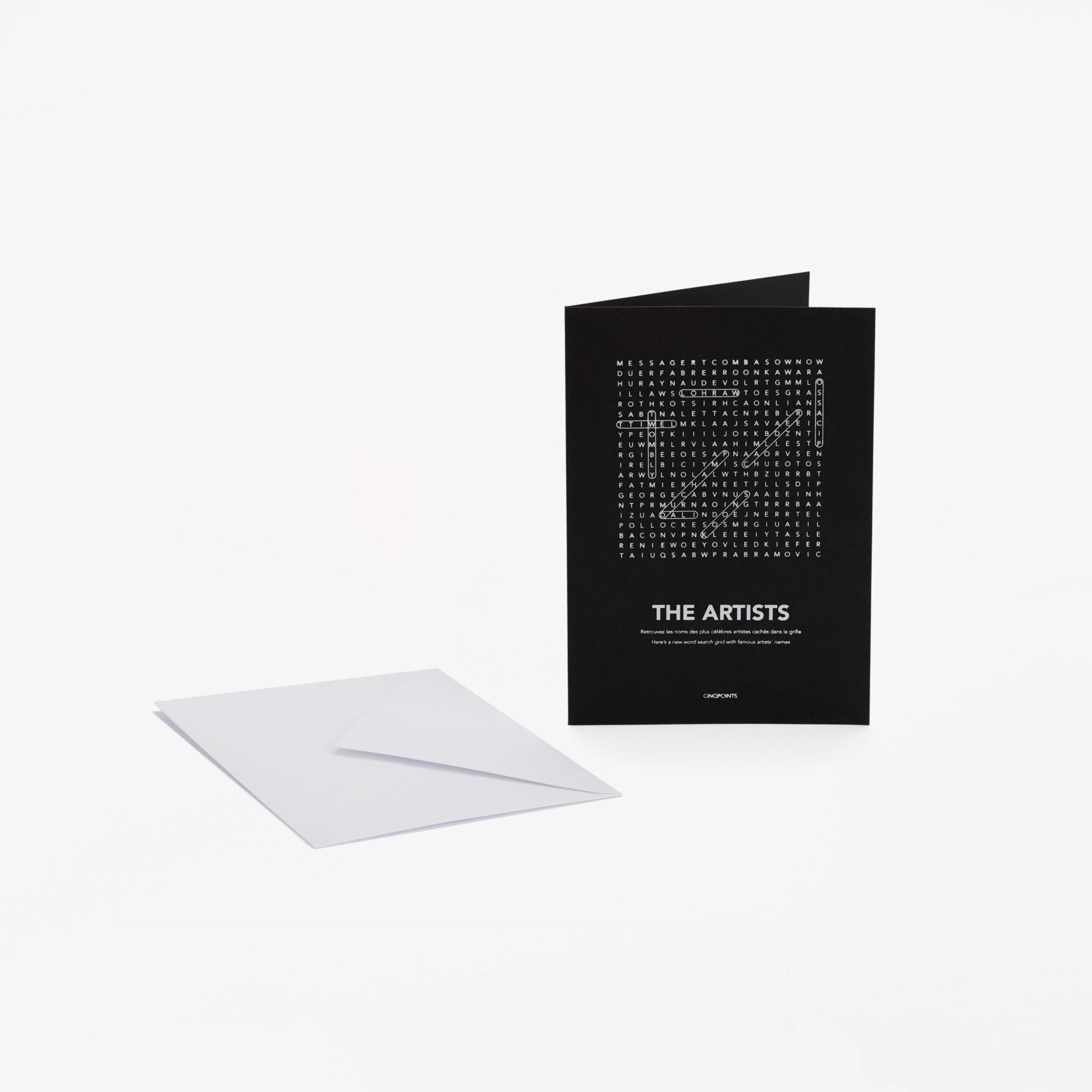 A black greeting card with white text "The Artists" and a word-search style crossword, shown standing upright next to a white envelope on a light gray background.