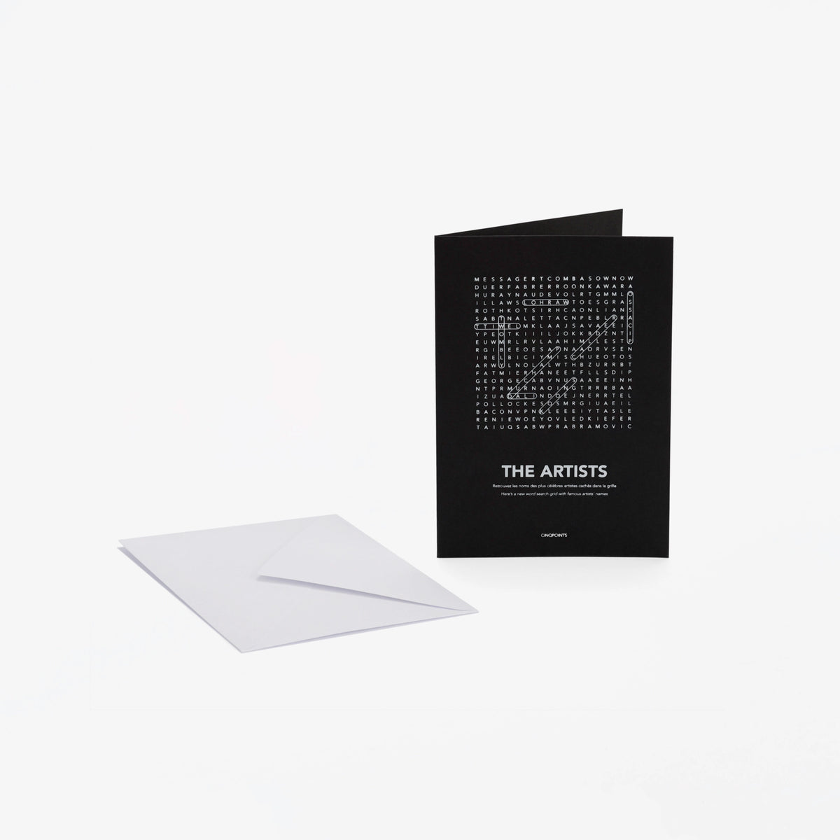 A black greeting card with white text "The Artists" and a word-search style crossword, shown standing upright next to a white envelope on a light gray background.