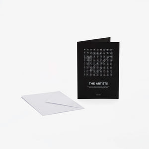 A black greeting card with white text "The Artists" and a word-search style crossword, shown standing upright next to a white envelope on a light gray background.