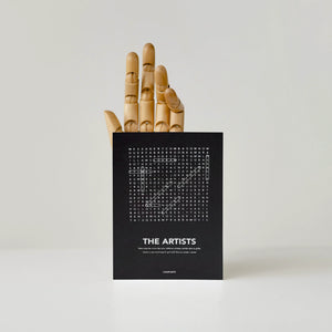 A black greeting card with white text "The Photographers" and a word-search style crossword, shown standing upright in front of a wooden hand behind it against a light gray background.