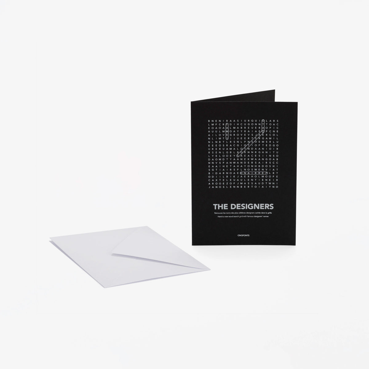 A black greeting card with white text "The Designers" and a word-search style crossword, shown standing upright next to a white envelope on a light gray background.