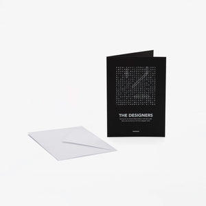 A black greeting card with white text "The Designers" and a word-search style crossword, shown standing upright next to a white envelope on a light gray background.