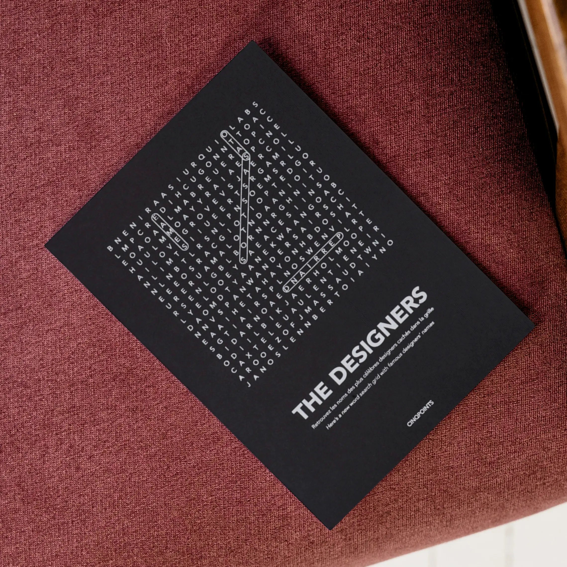 A black greeting card with white text "The Designers" and a word-search style crossword, lies diagonally on a textured burgundy fabric and wooden chair, shown from above.