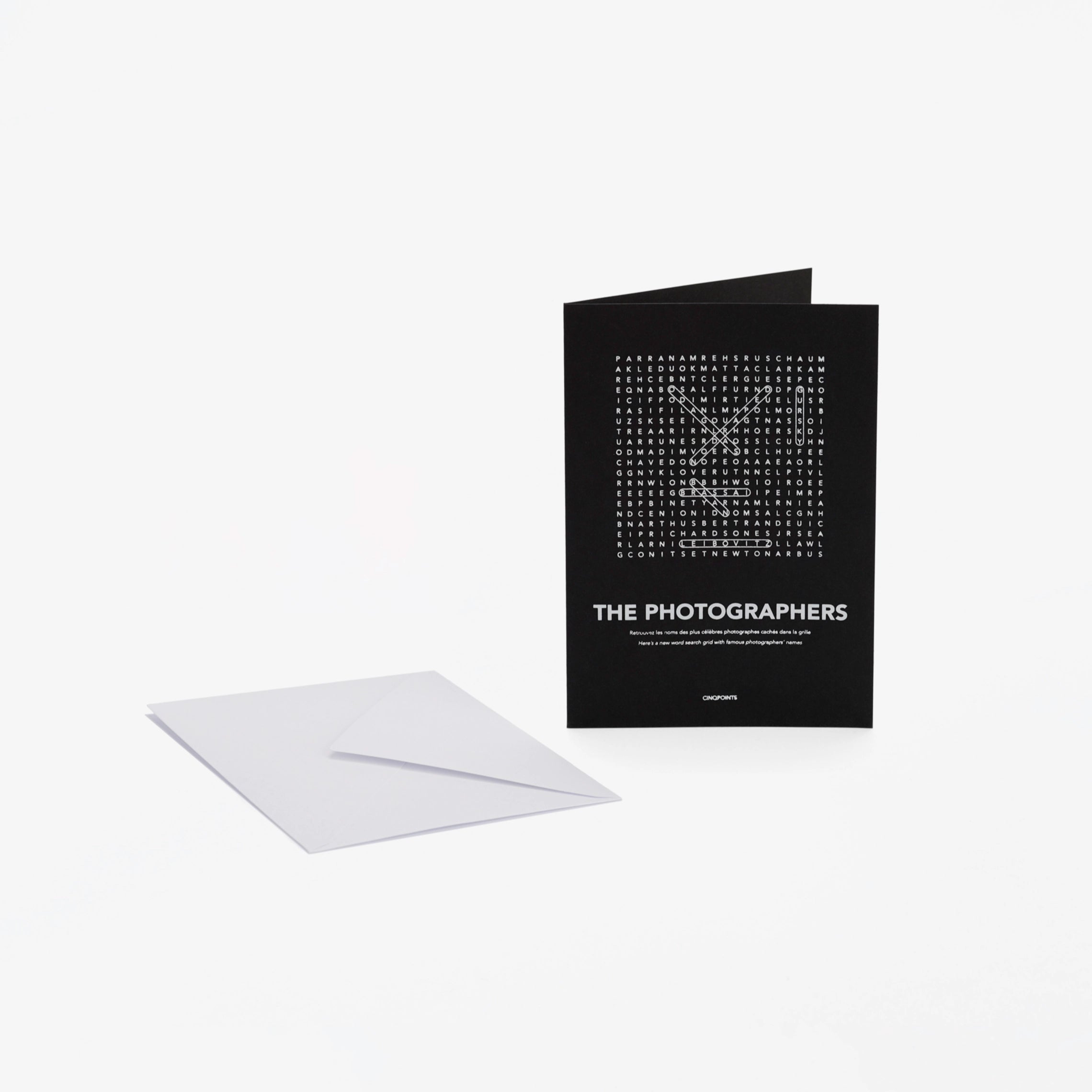 A black greeting card with white text "The Photographers" and a word-search style crossword, shown standing upright next to a white envelope on a light gray background.