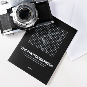 A black greeting card with white text "The Photographers" with a white word-search style crossword, styled on a white surface with a vintage camera and white envelopes nearby on a white table.