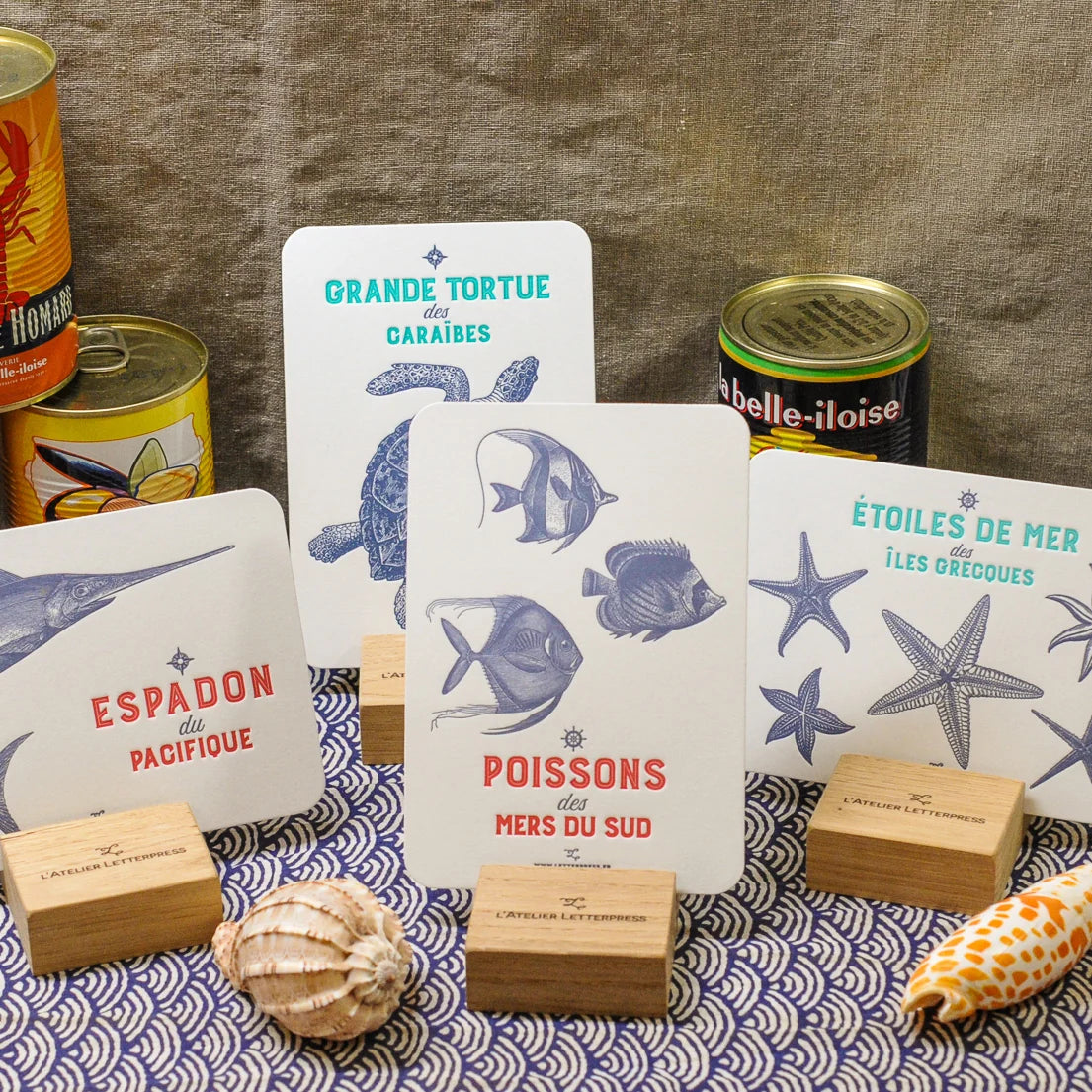 A set of French text set of 10 Maritime sea creatures art print cards displayed in wooden stands on a patterned blue and white tablecloth seashells and seafood can. The cards are coloured with red and gray text and illustrations.
