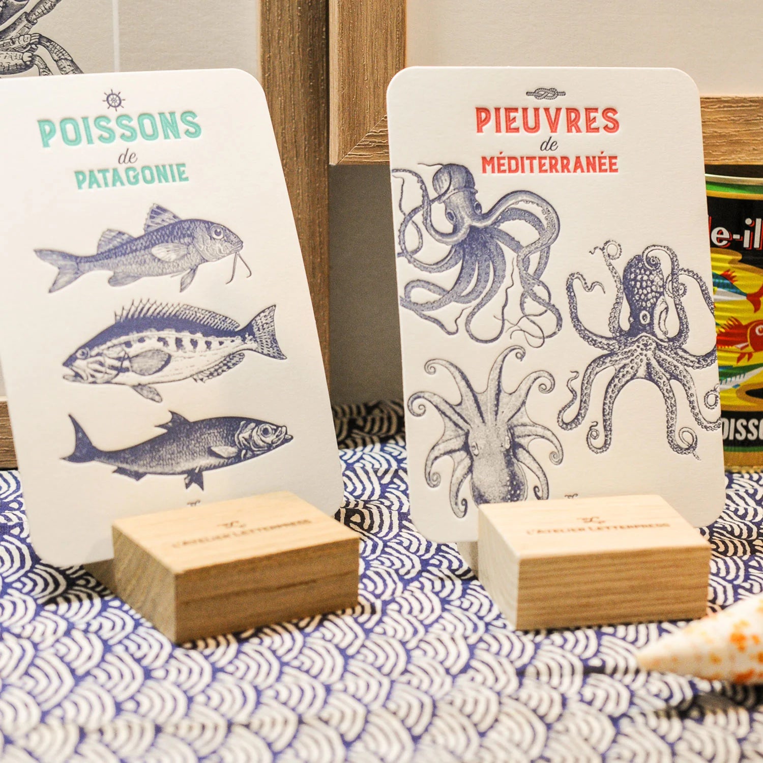 Two art print cards displayed on small wooden stands on a blue-and-white patterned surface. The left card has 3 fish illustrations, and the right card has 3  octopuses illustrations, each with text.