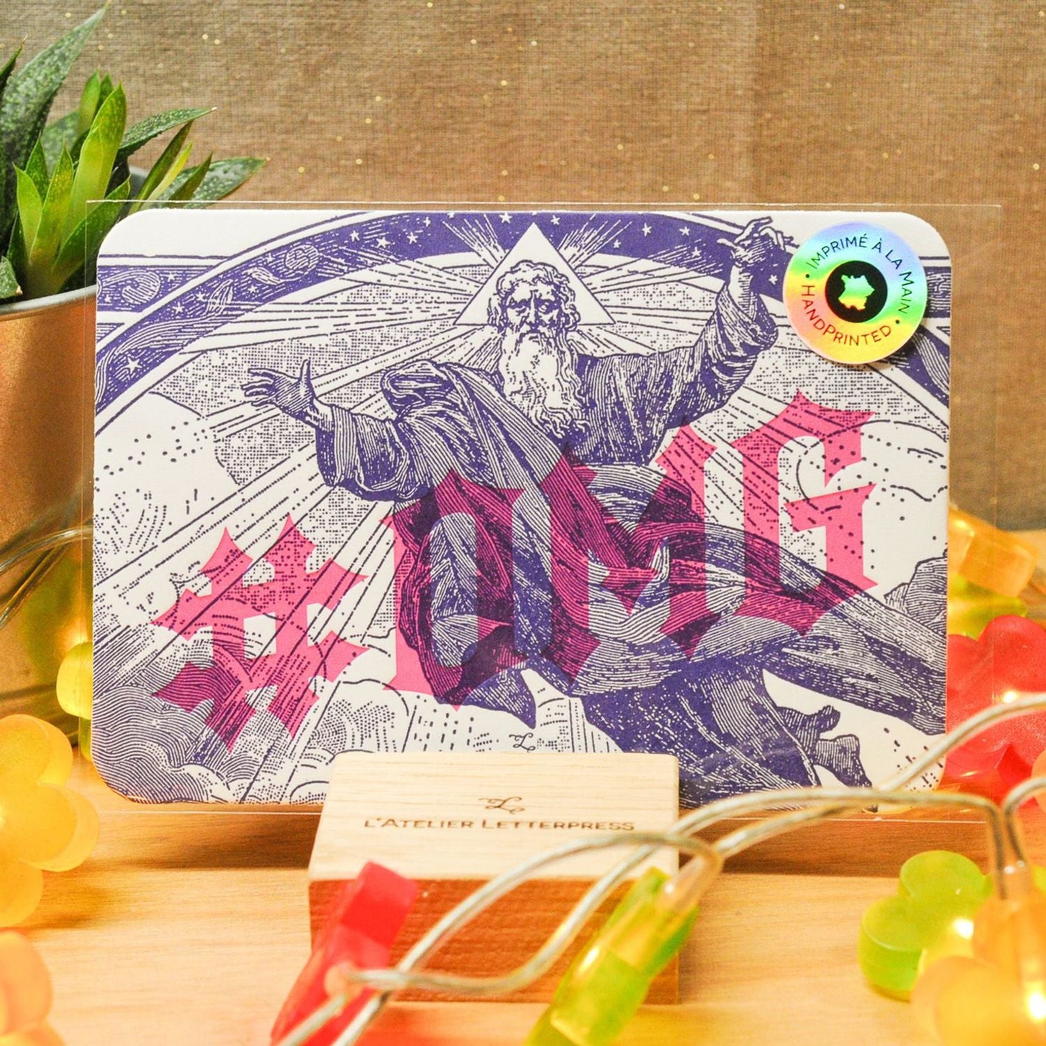 An A6 card with rounded corners with '#OMG' text and a vintage-style illustration of a bearded man, displayed on a wooden stand with colourful fairy lights and a burlap material backdrop.