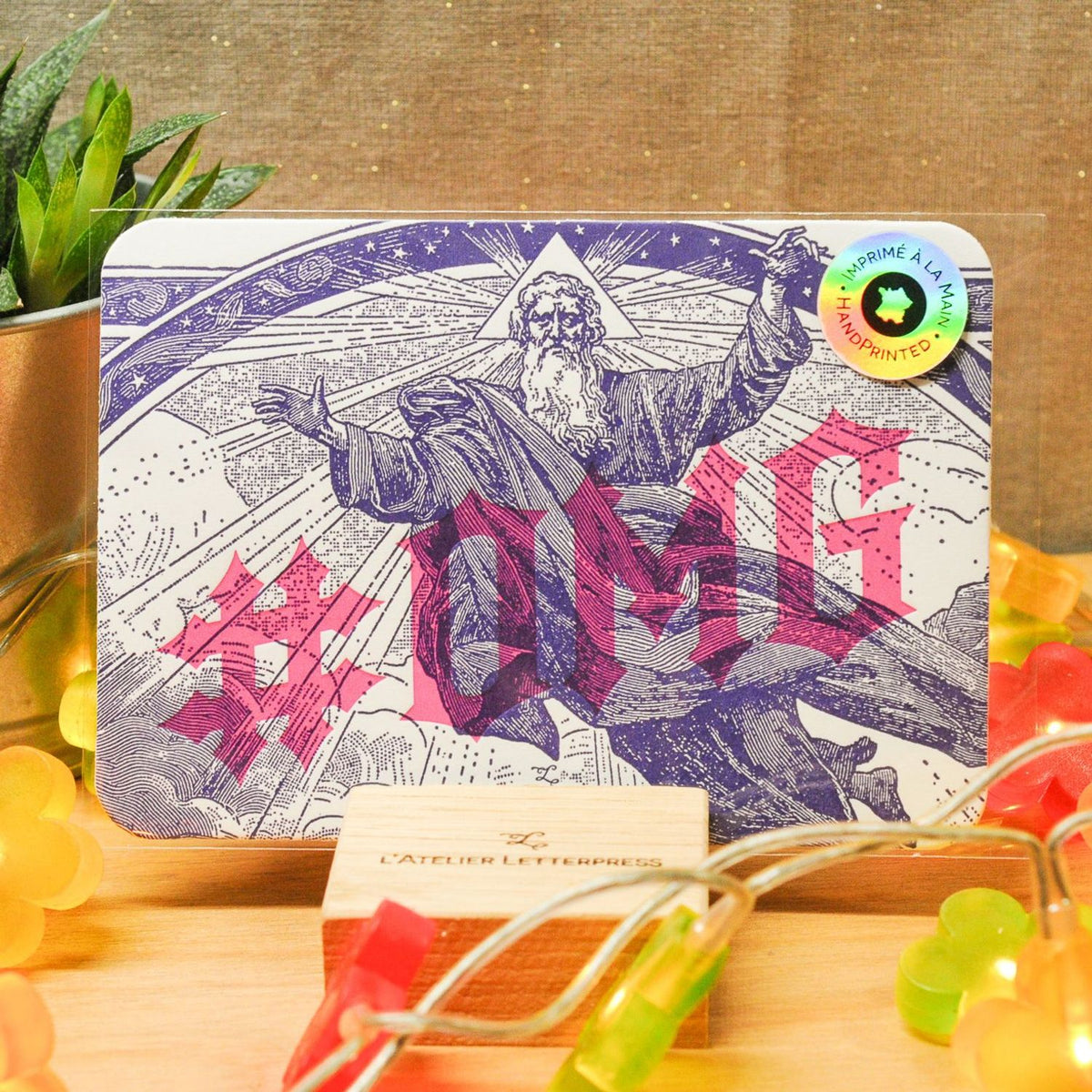 An A6 card with rounded corners with '#OMG' text and a vintage-style illustration of a bearded man, displayed on a wooden stand with colourful fairy lights and a burlap material backdrop.