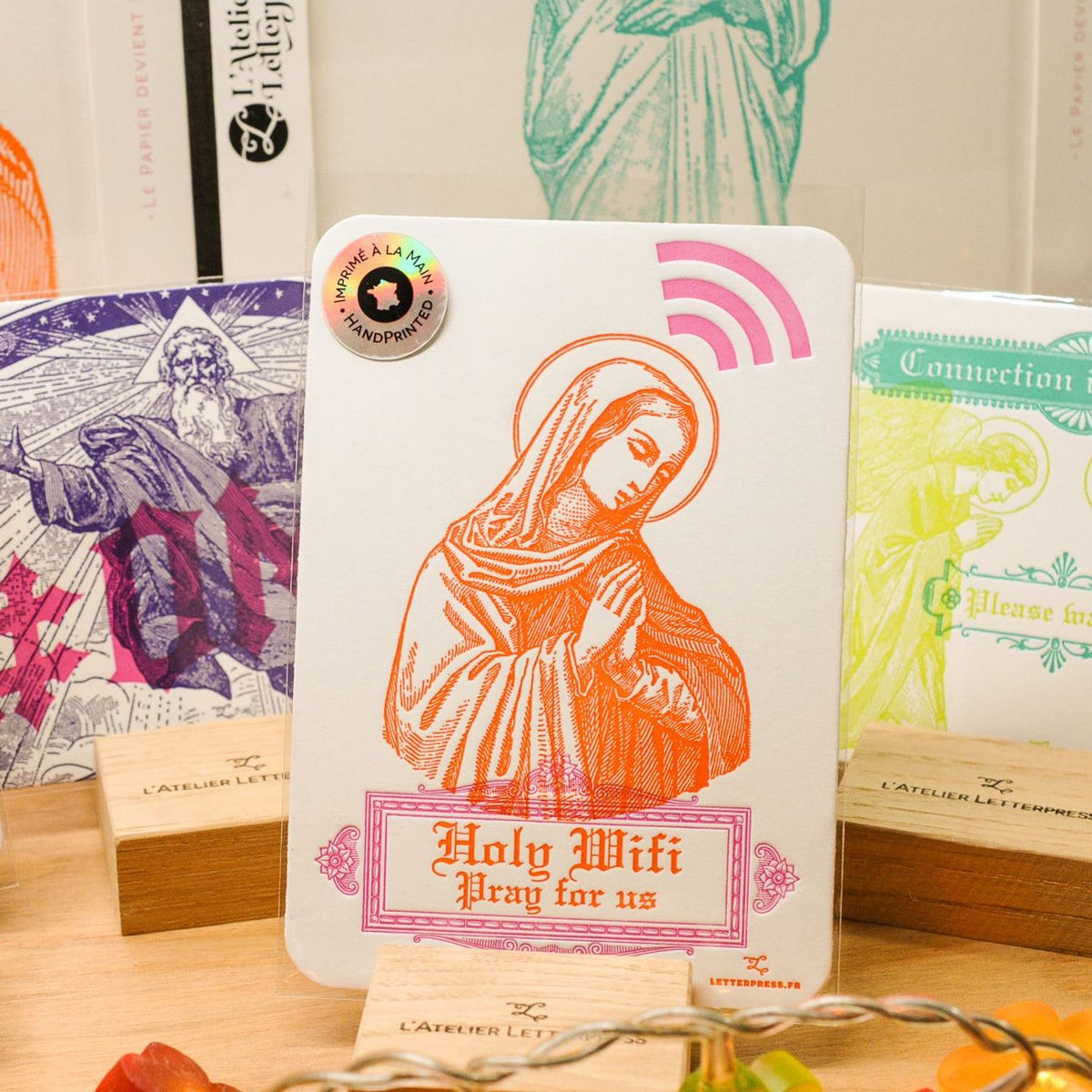A white rectangular art print card featuring an orange line drawing of Mary with the text 'Holy Wifi Pray for us' and a pink wireless symbol, displayed on a wooden surface with other devotional cards in the background.