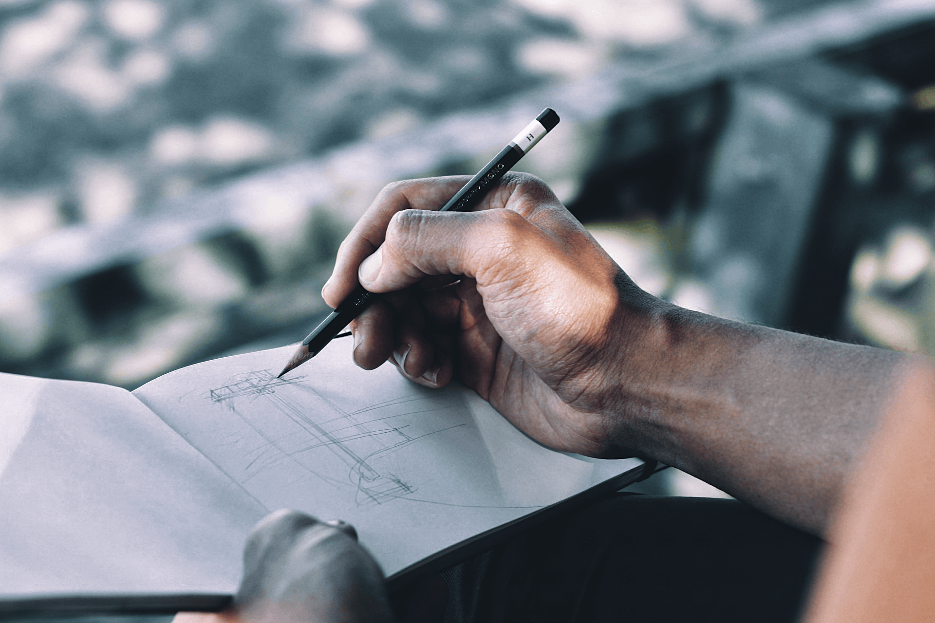 Hand holding a pencil over a notebook with a blurred outdoor background