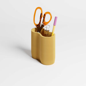 A ribbed mustard yellow pen holder or desk organiser on a light gray background, containing scissors, a black binder clip, and a yellow pen.