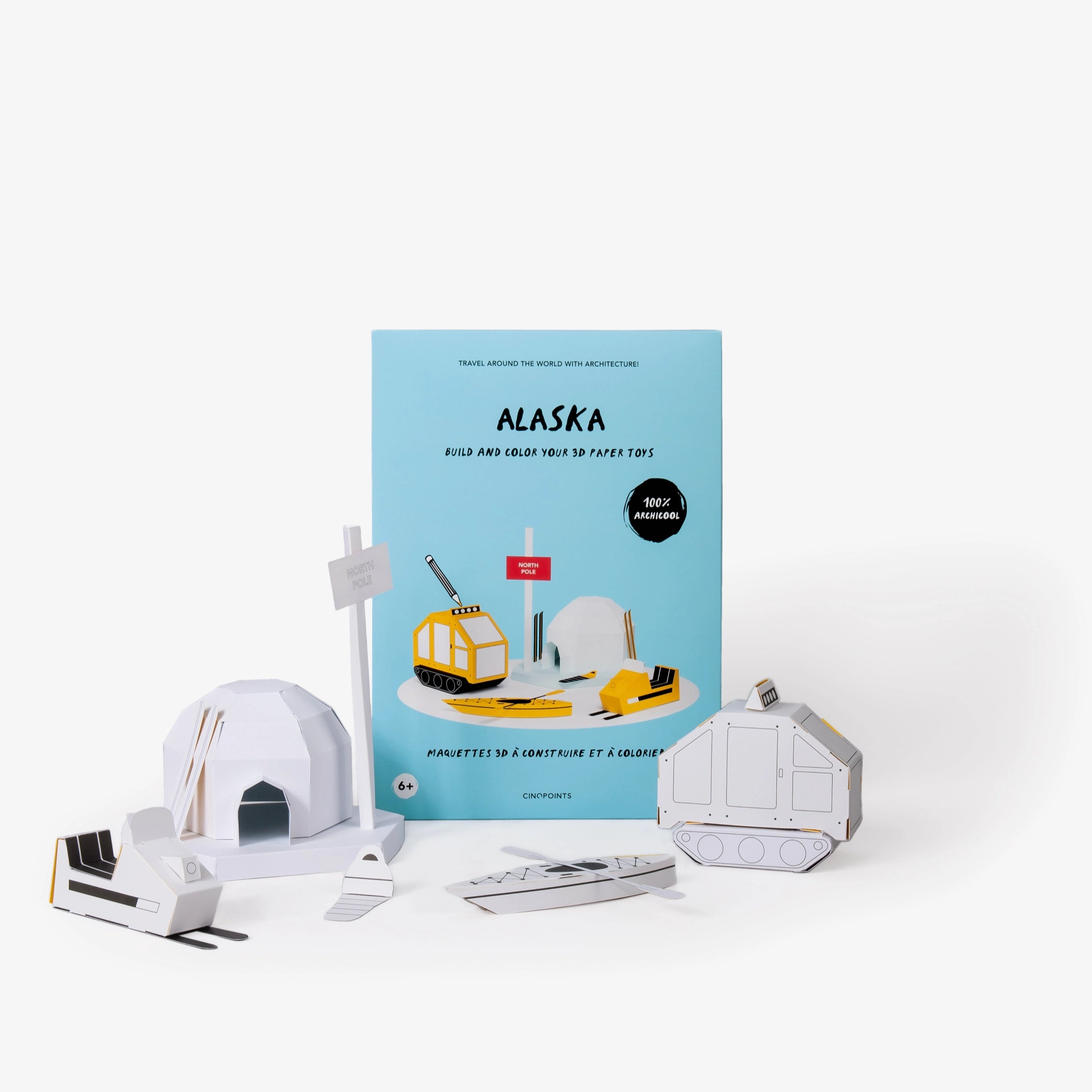Alaska paper model kit folder featuring a light blue cover standing upright behind  several white assembled paper craft models including a north pole sign, igloo, snowmobile and other winter items against a clean white background.
