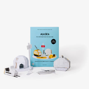 Alaska paper model kit folder featuring a light blue cover standing upright behind  several white assembled paper craft models including a north pole sign, igloo, snowmobile and other winter items against a clean white background.
