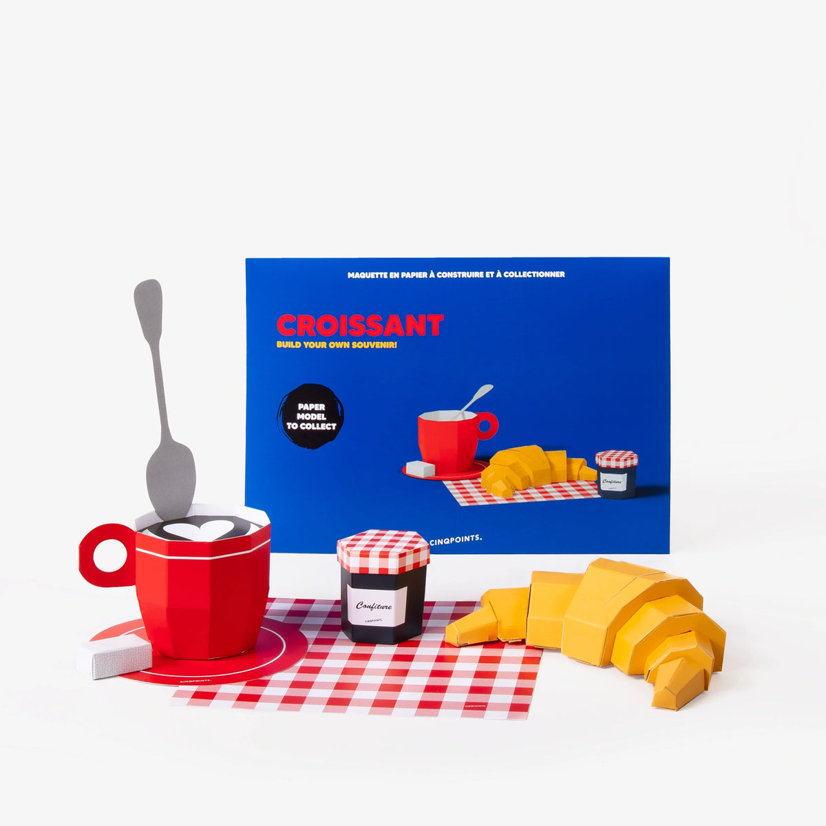 A blue collectible paper model kit envelope behind, and in front paper models of a red mug, croissant, a jam jar, and a red-and-white checkered napkin on a white background.