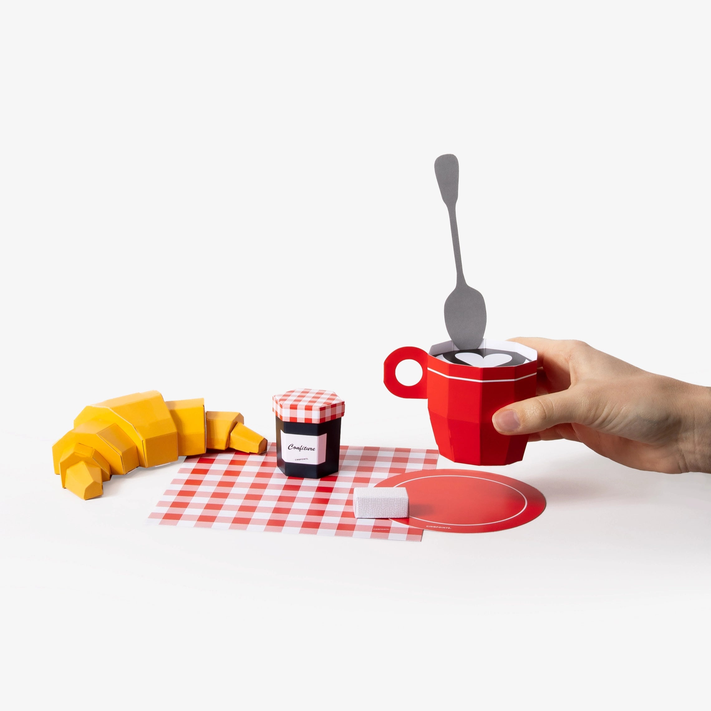 A hand holds a red paper model coffee mug with a spoon inside, alongside paper models of a croissant, a jam jar, a red-and-white checkered napkin, and a red coffee coaster on a white background.