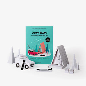 Mont Blanc paper model kit folder displayed on a white background, and in front a red and white camper van, a wooden A-frame cabin, chairs, trees, and other winter-themed accessories, all still white but assembled.